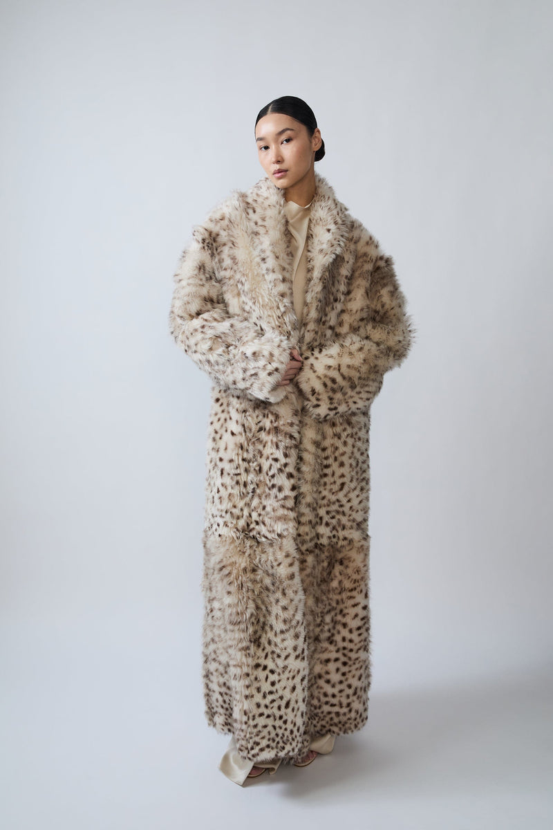 A model wearing a long, plush faux fur coat with a beige and brown animal print pattern, standing against a plain light gray studio background.