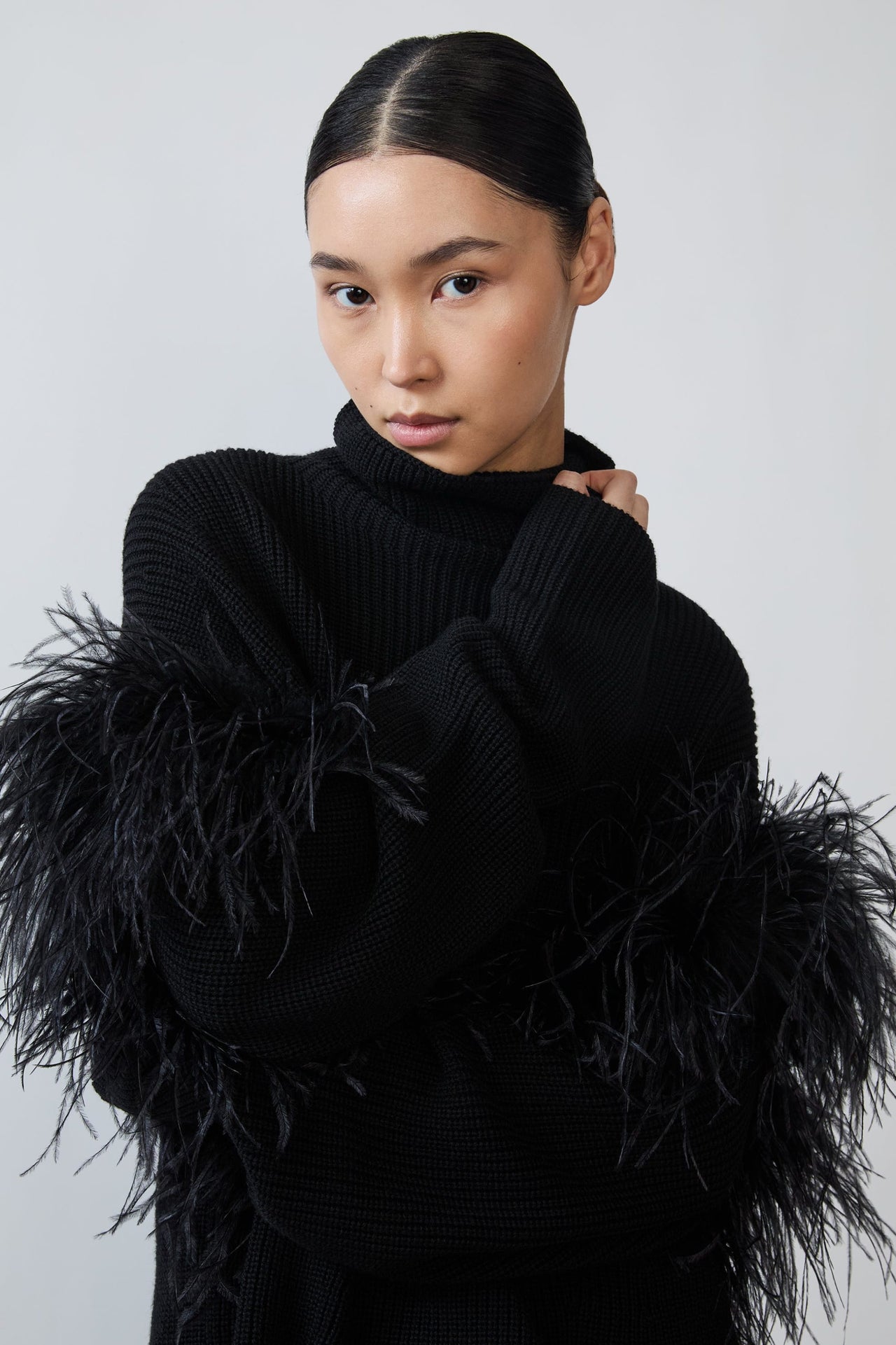A model wearing a black ribbed turtleneck sweater with dramatic black feather trim on the sleeves, posed against a light gray studio background.