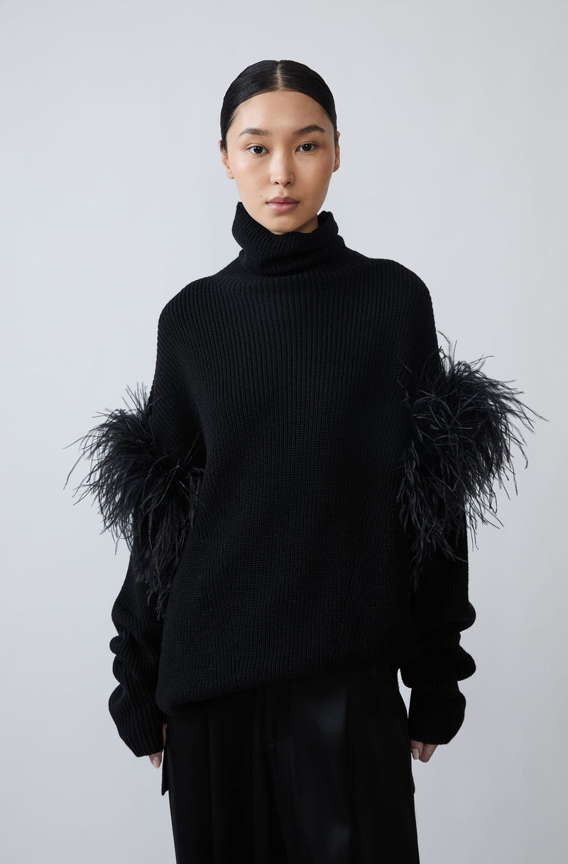 A model wearing a black ribbed turtleneck sweater with dramatic black feathered shoulder details, paired with black trousers, against a plain light gray studio background.