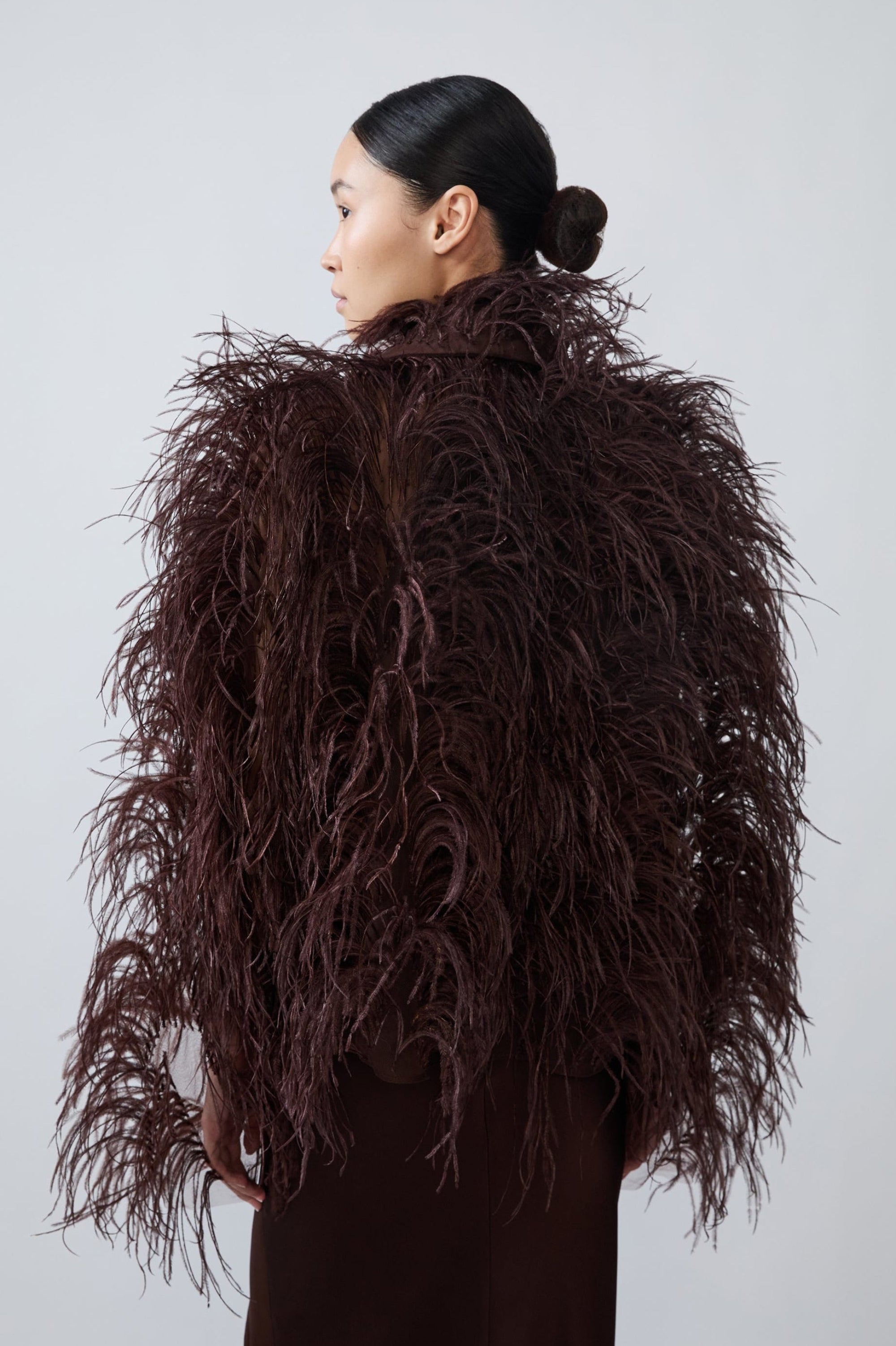 A model shown from the back wearing a voluminous, dark brown feathered jacket or coat over a matching dark brown garment, set against a plain light gray studio background.