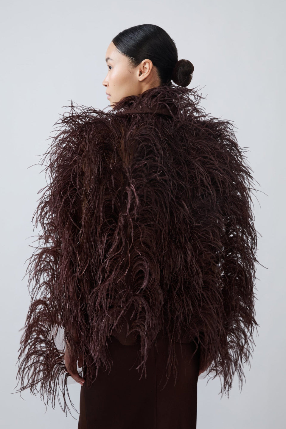A model shown from the back wearing a voluminous, dark brown feathered jacket or coat over a matching dark brown garment, set against a plain light gray studio background.