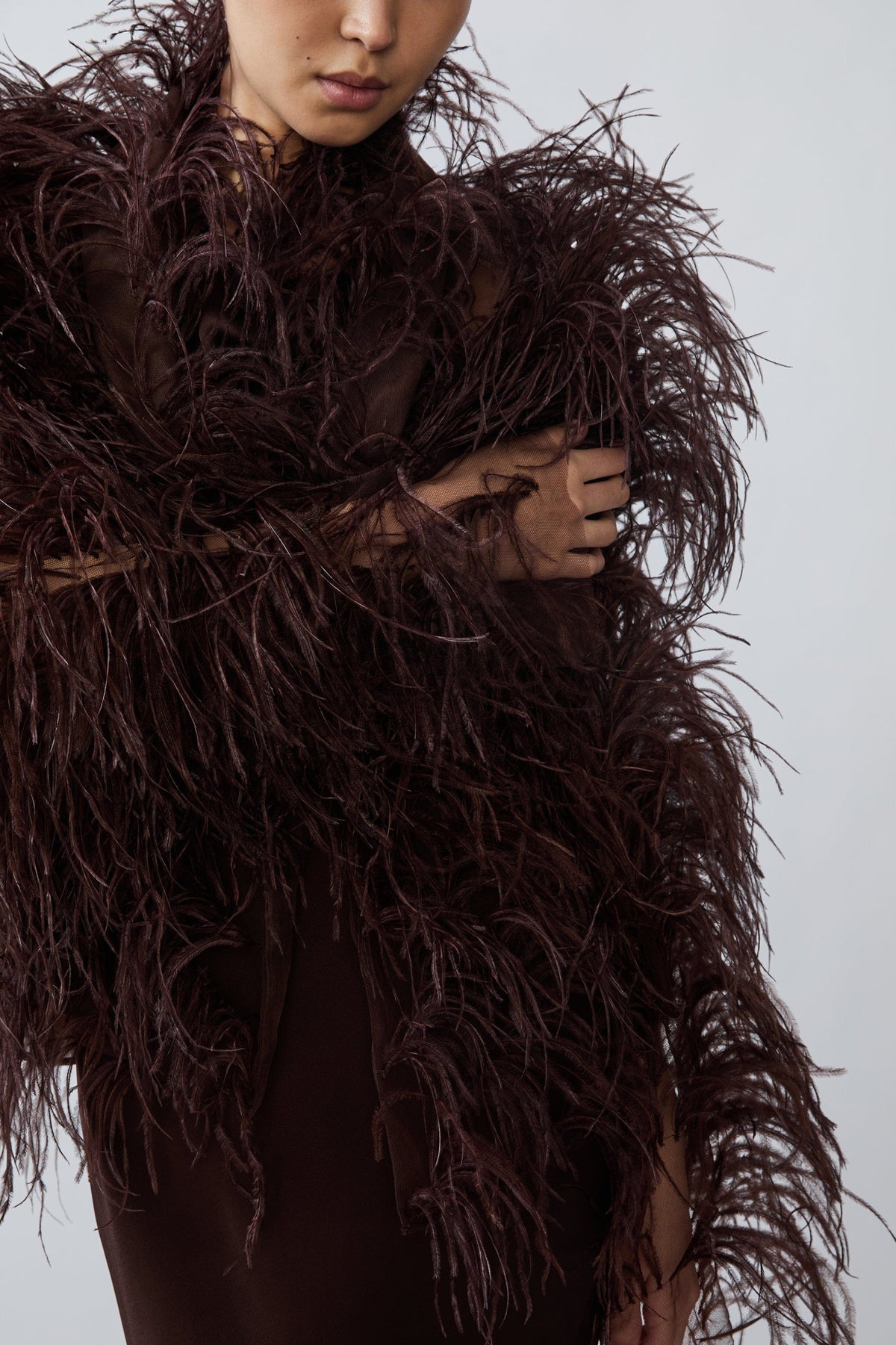 A close-up of a model wearing a dark brown, voluminous feathered garment or accessory over a matching dark brown top, set against a plain light gray background.