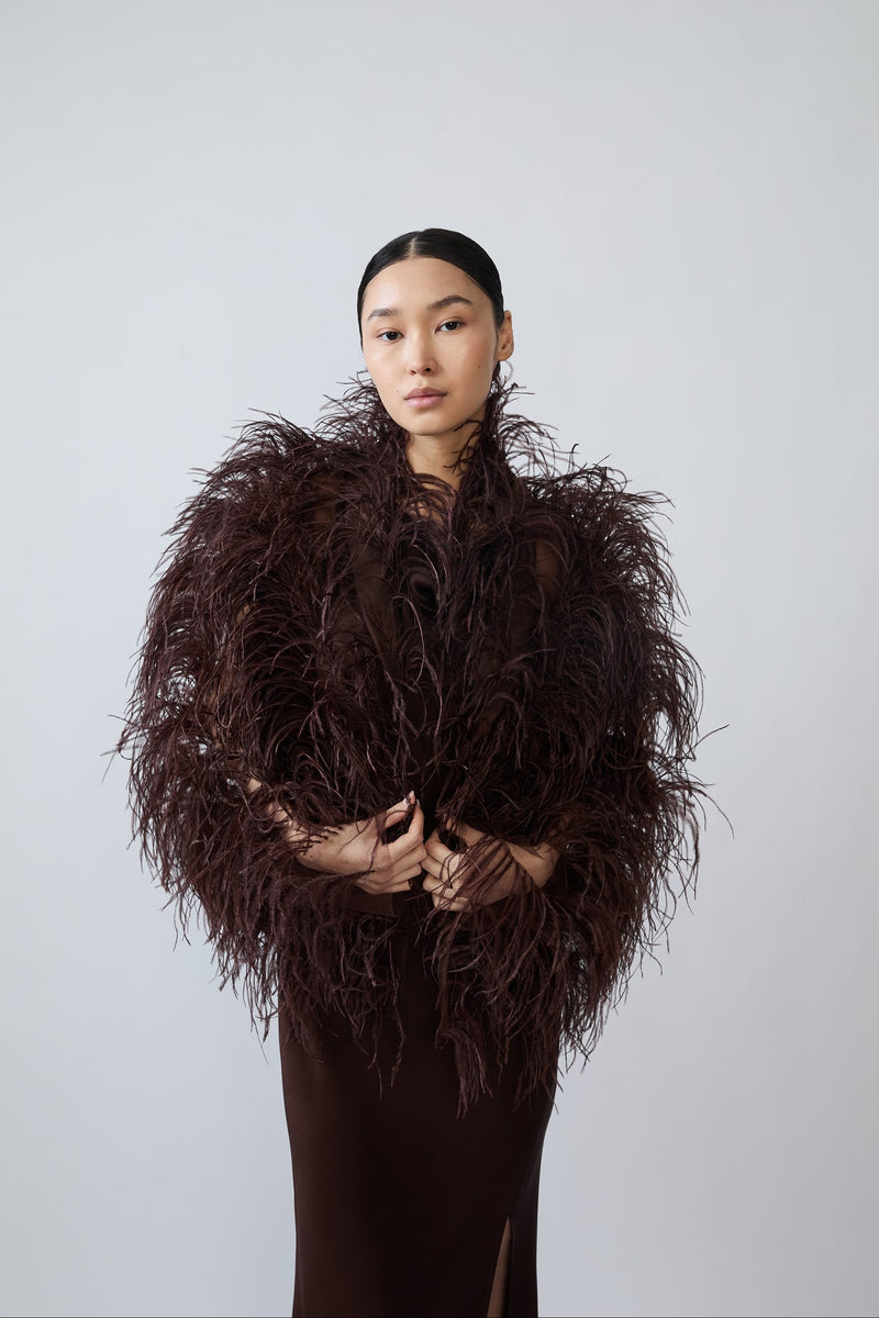 A model wearing a dark brown, voluminous feathered jacket over a matching dark brown dress, standing against a solid light gray background.