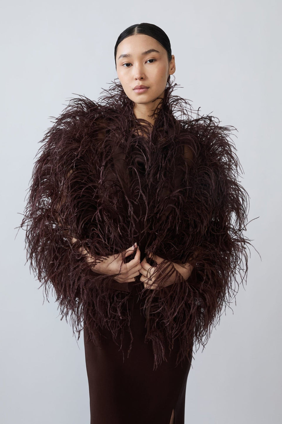 A model wearing a dark brown, voluminous feathered jacket over a matching dark brown dress, standing against a solid light gray background.