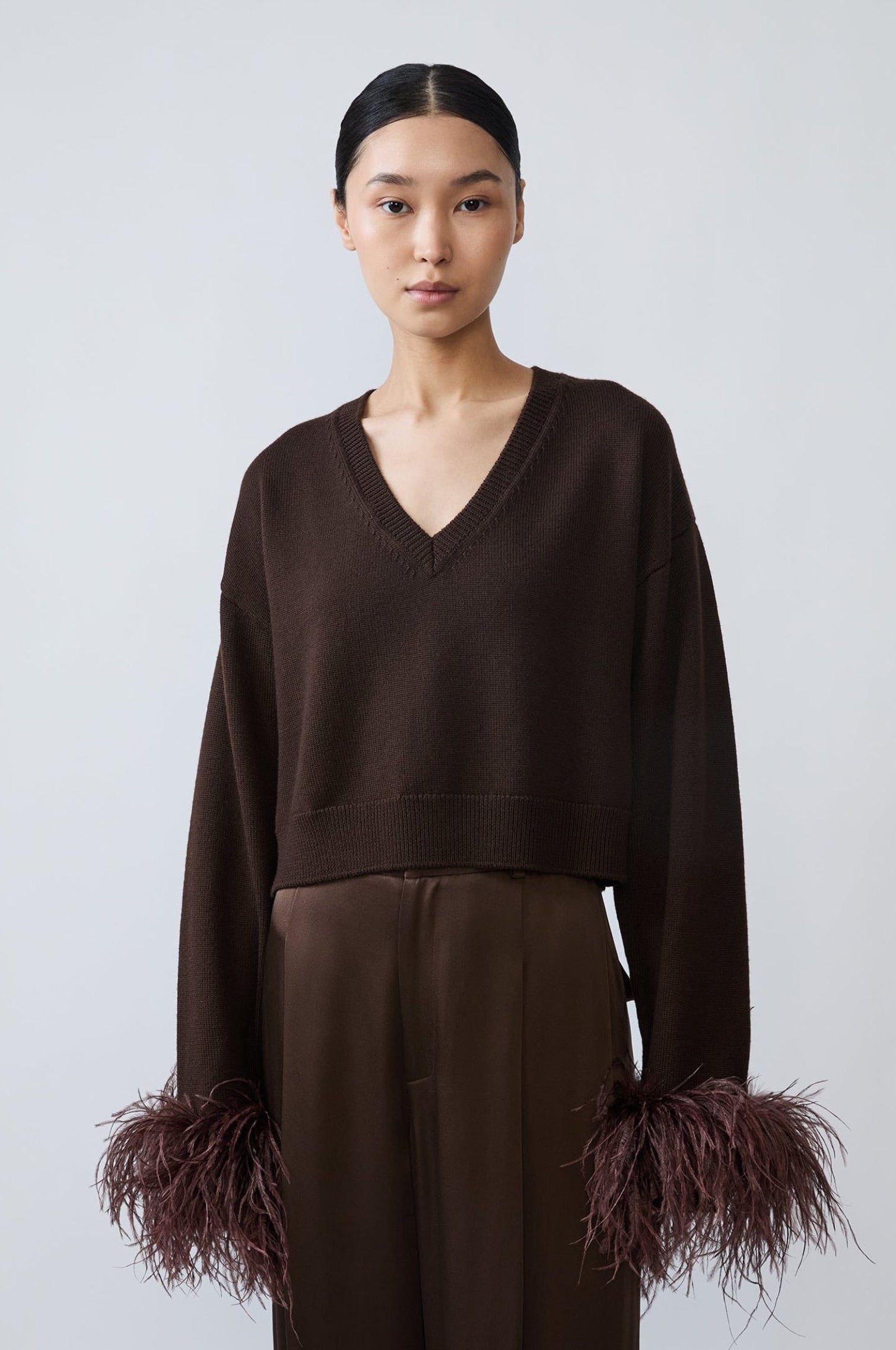 A model stands against a light gray studio background wearing a dark brown cropped V-neck sweater with feathered cuffs and matching dark brown wide-leg trousers.