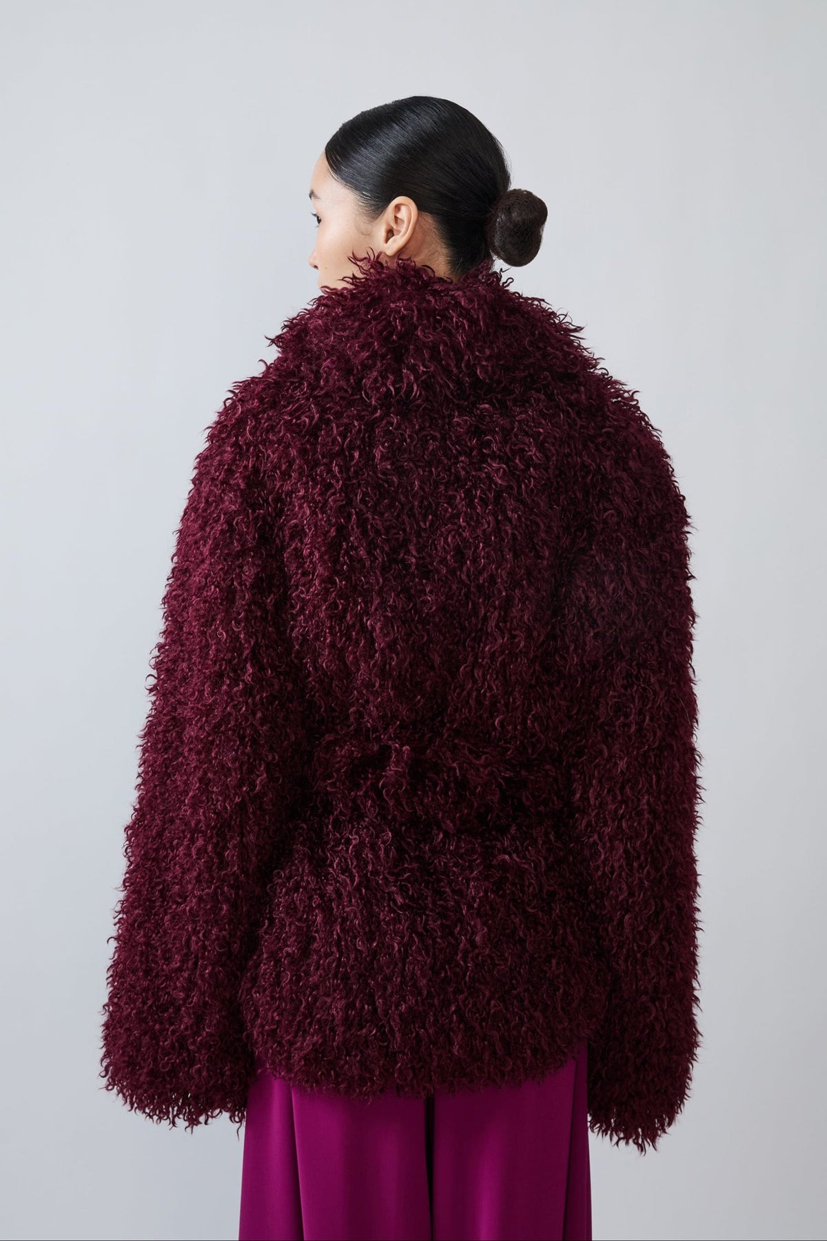 A model shown from the back wearing a shaggy, textured burgundy wrap jacket and magenta trousers against a plain light gray studio background.