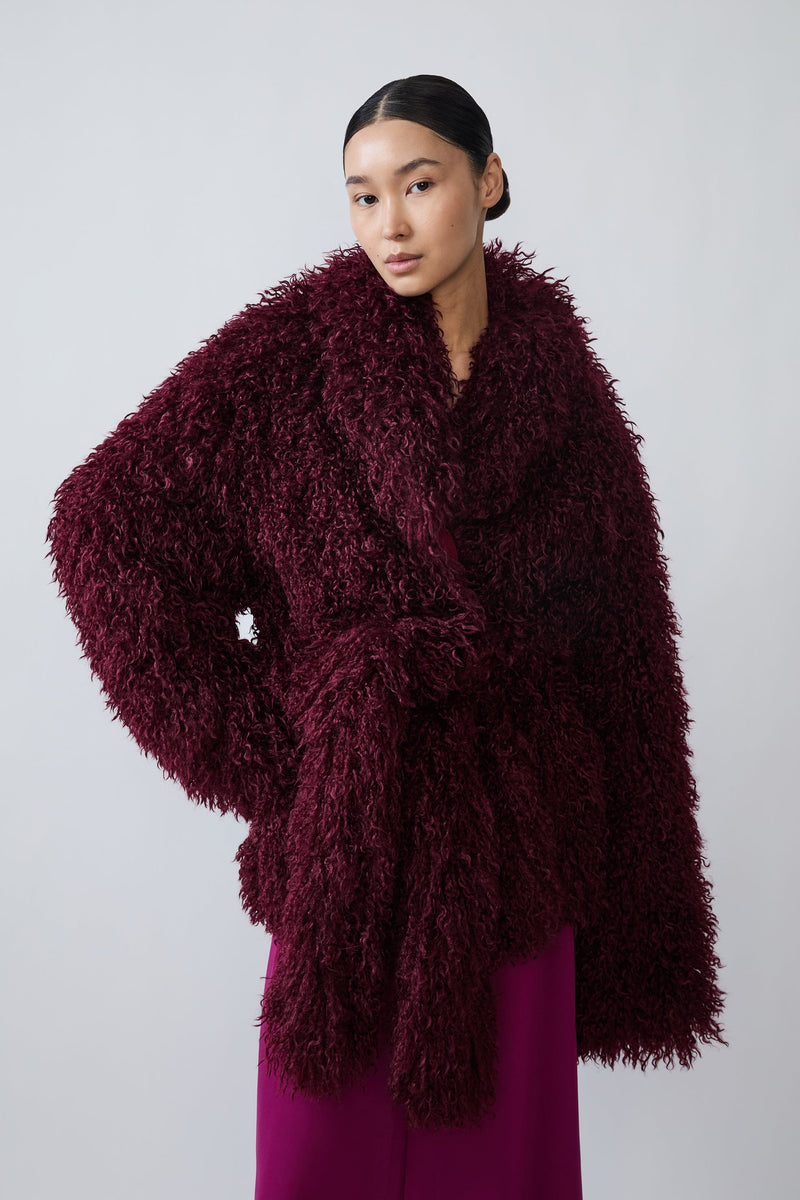 A model wearing a voluminous, deep burgundy shaggy faux-fur wrap jacket over a magenta dress, photographed against a neutral light gray studio background.
