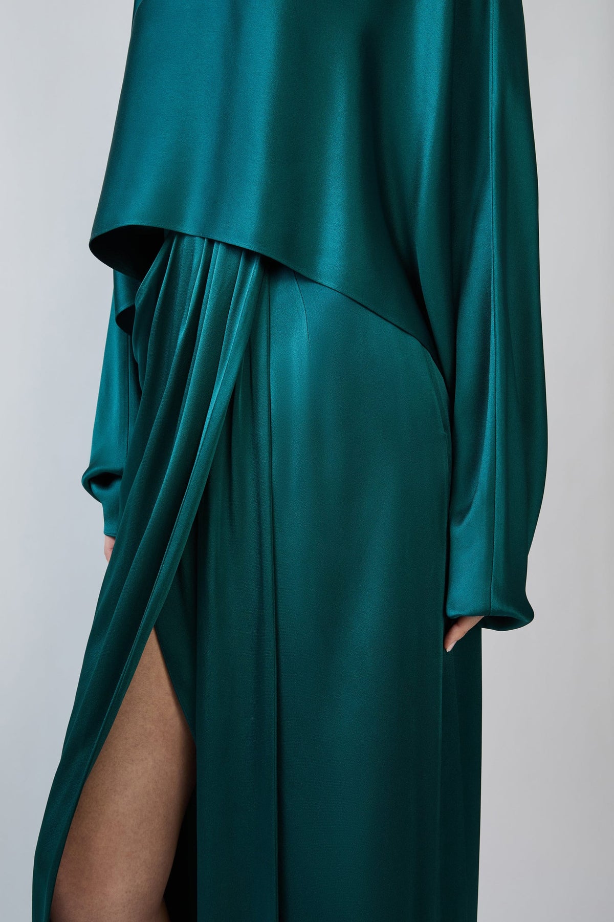 A close-up of a long flowing satin dress in deep teal green, featuring draped layers, a high side slit, and long sleeves, shown on a model against a light gray background.