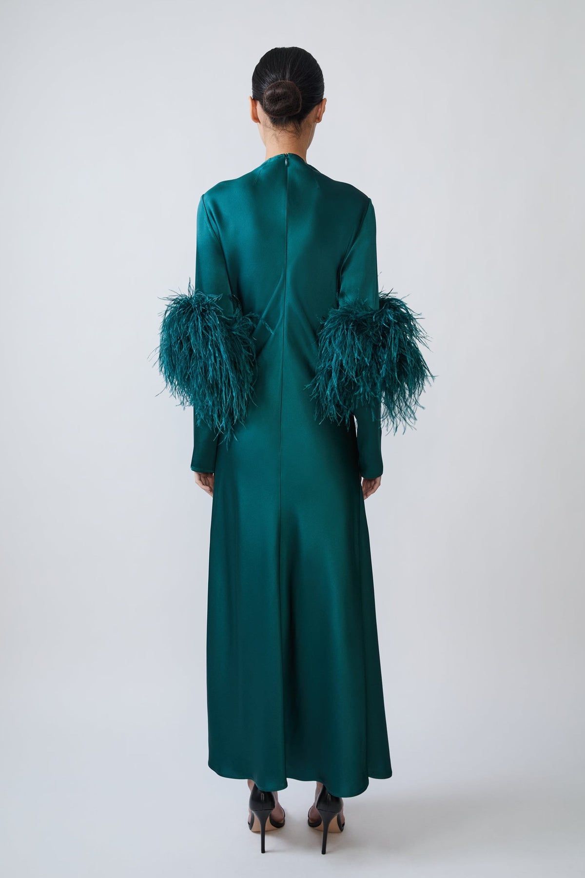 A model viewed from the back wearing a long emerald green satin dress with dramatic feathered sleeves, set against a plain light gray studio background.