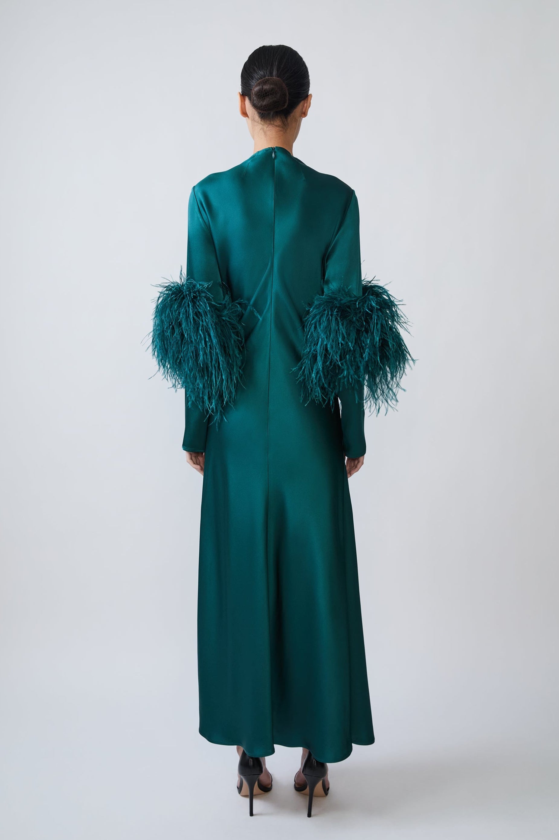 A model viewed from the back wearing a long emerald green satin dress with dramatic feathered sleeves, set against a plain light gray studio background.