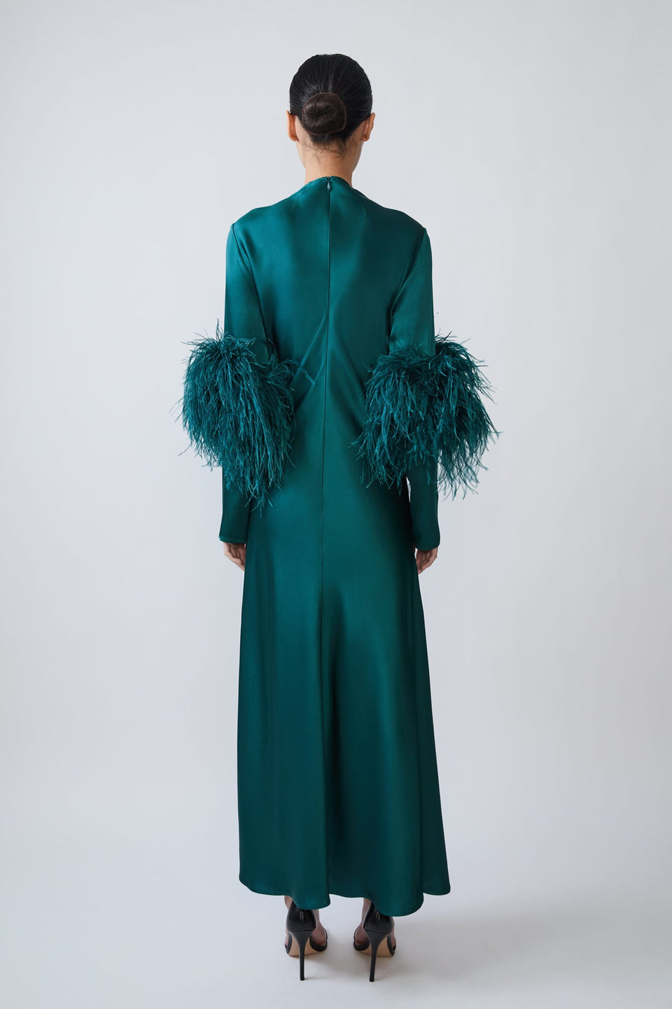 A model viewed from the back wearing a long emerald green satin dress with dramatic feathered sleeves, set against a plain light gray studio background.