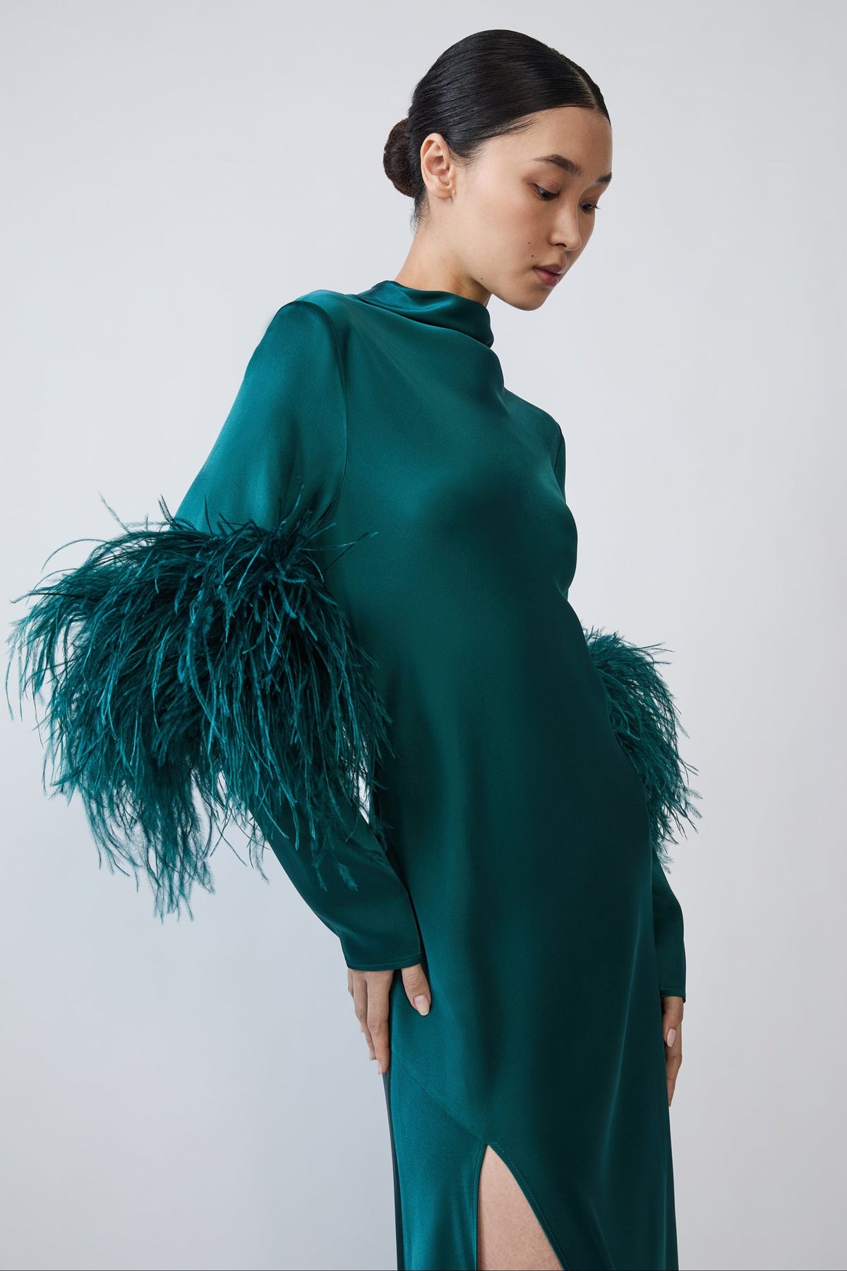 A model wearing a dark teal green satin dress with dramatic feathered sleeves, photographed against a neutral light gray studio background.