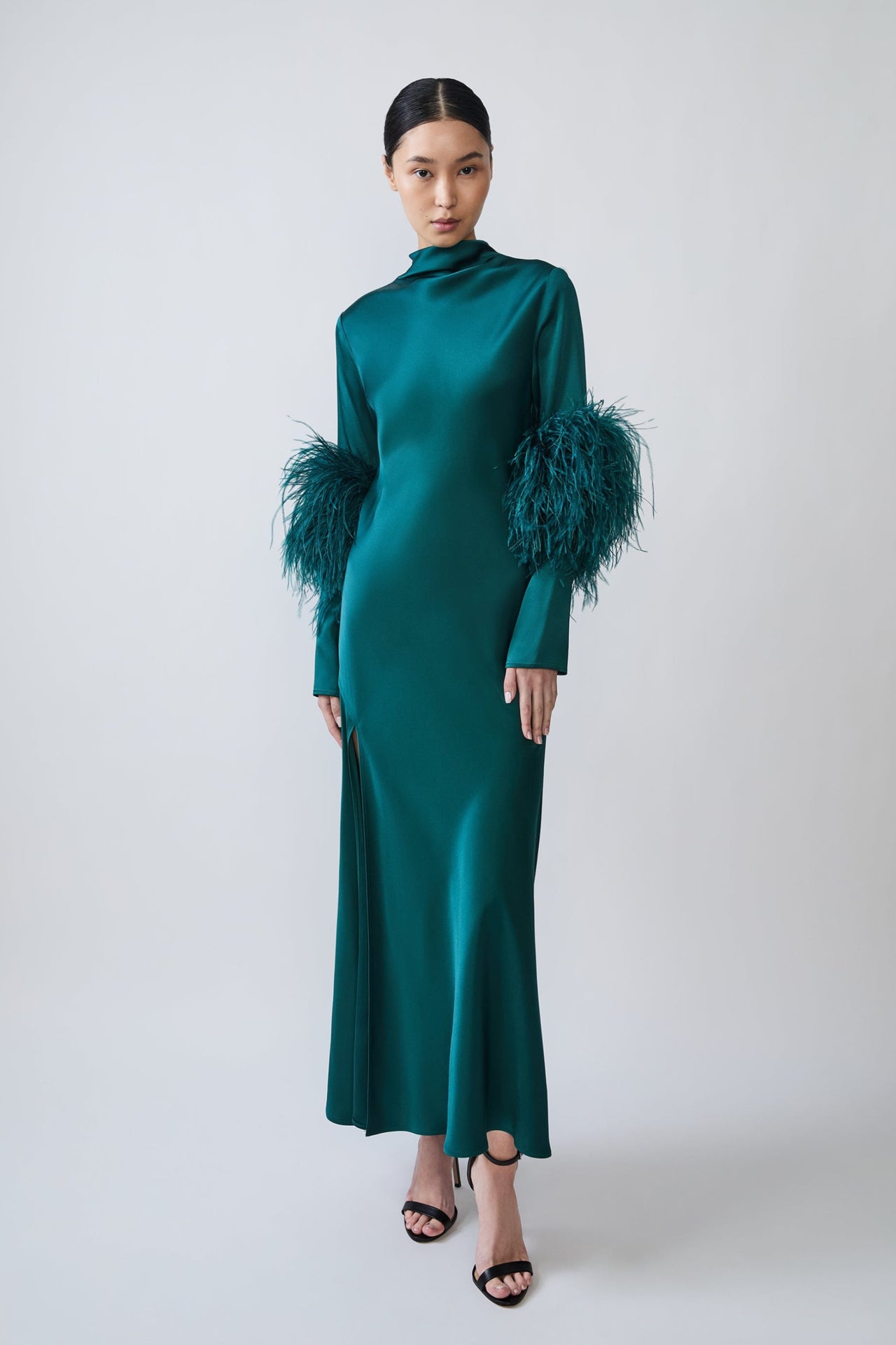 A model wearing a floor-length emerald green satin gown with high ruffled mock necklines and dramatic feathered sleeves, paired with black strappy heels, against a light gray studio background.