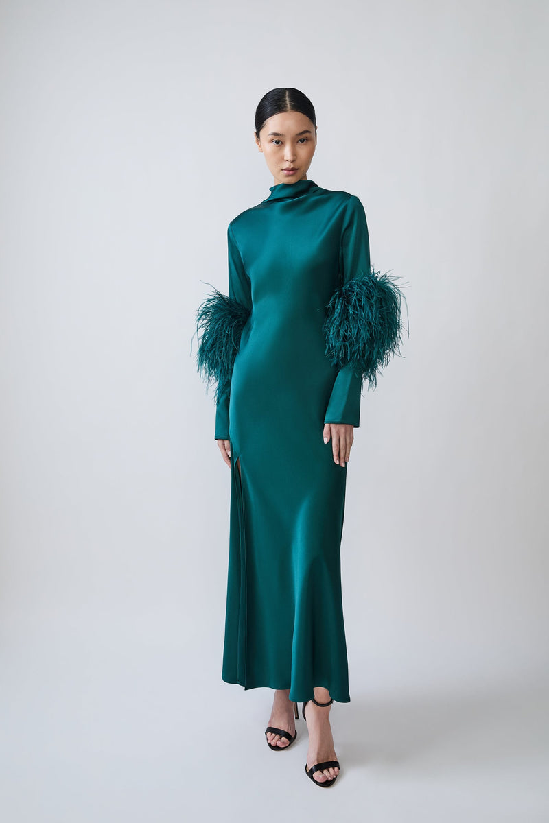 A model wearing a floor-length emerald green satin gown with high ruffled mock necklines and dramatic feathered sleeves, paired with black strappy heels, against a light gray studio background.