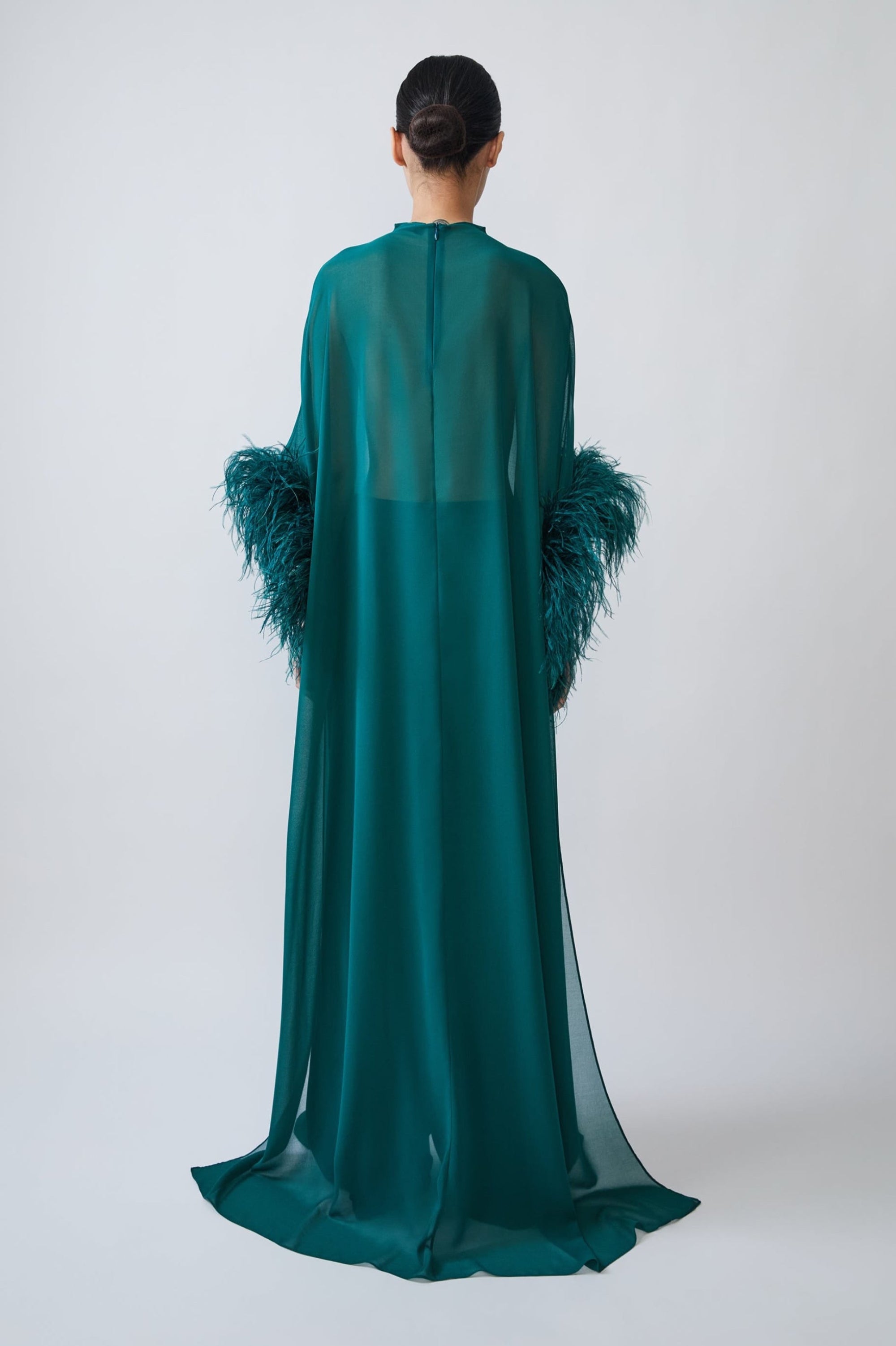 A model viewed from the back wearing a floor-length teal green sheer gown with dramatic feathered sleeves, set against a plain light gray studio background.