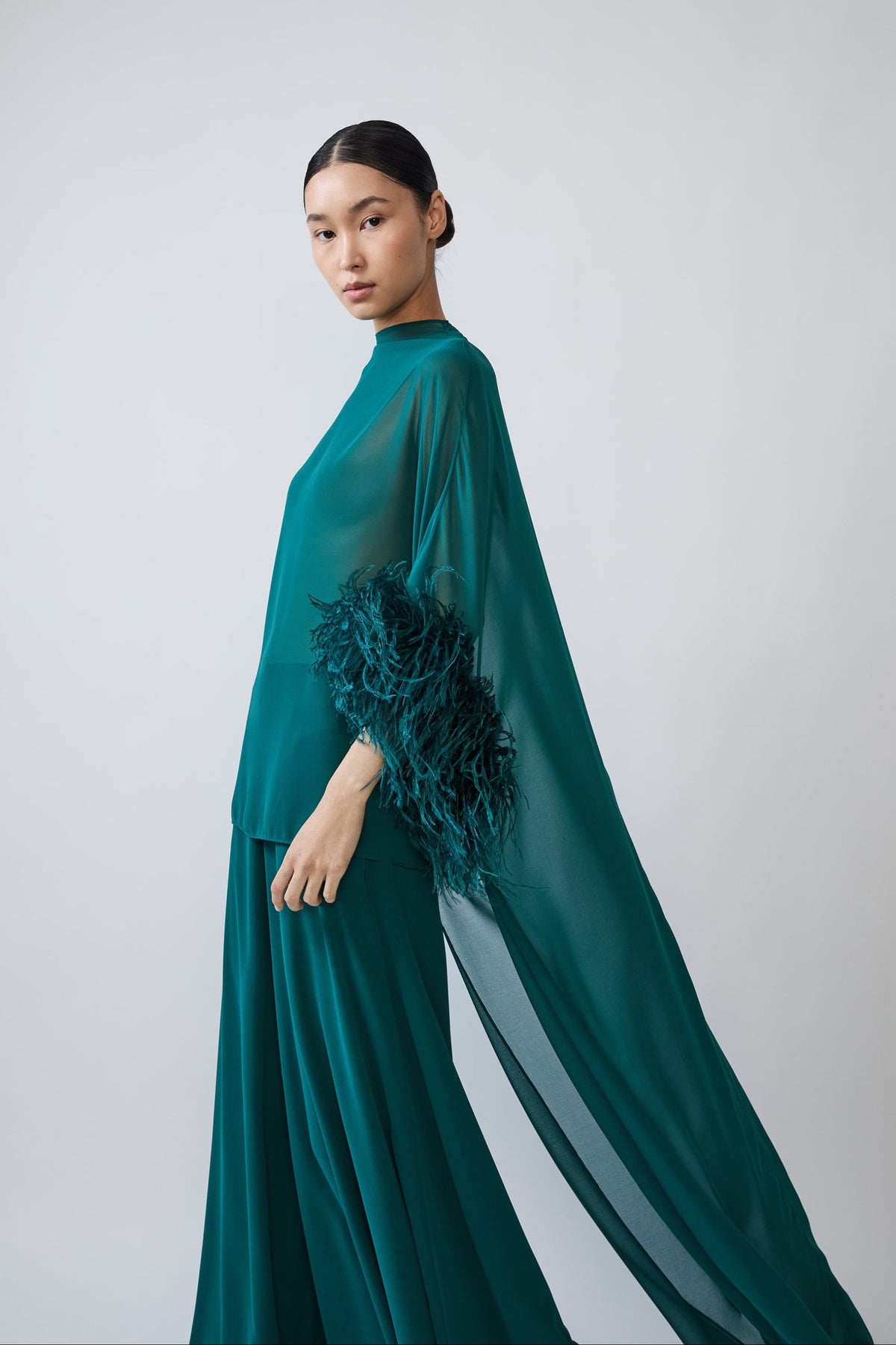 A model wearing a floor-length teal green gown with a sheer cape-like sleeve and feathered detailing on the arm, photographed against a light gray studio background.