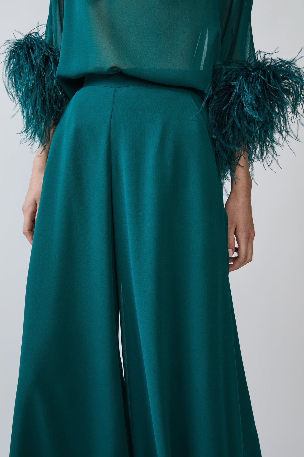A close-up of a model wearing high-waisted, wide-leg trousers in emerald green, paired with a matching sheer top featuring dramatic teal feathered cuffs, shot against a plain light gray background.