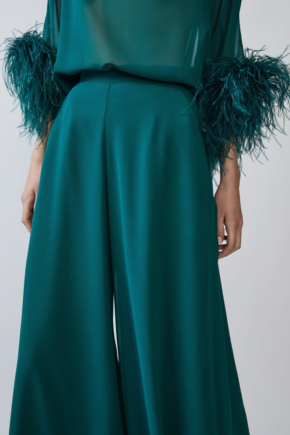A close-up of a model wearing high-waisted, wide-leg trousers in emerald green, paired with a matching sheer top featuring dramatic teal feathered cuffs, shot against a plain light gray background.