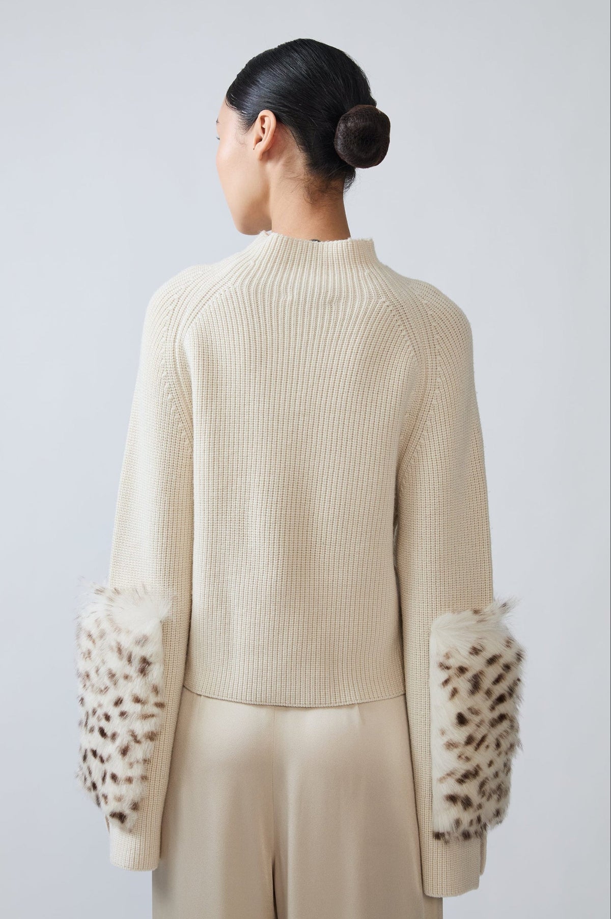 A model shown from the back wearing a cream ribbed knit sweater with leopard-print faux fur cuffs, paired with matching cream trousers, against a light gray studio background.
