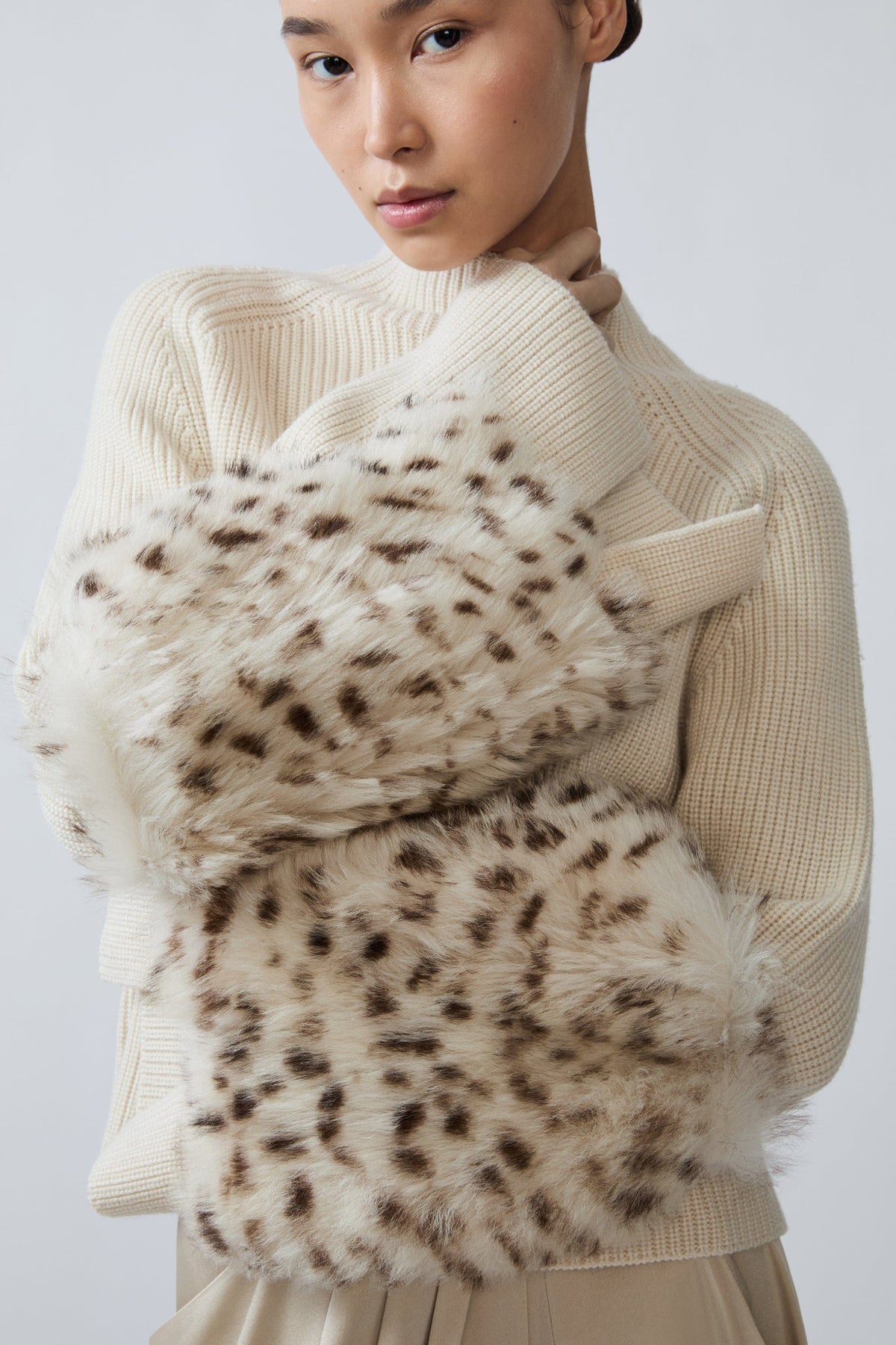 A model wearing a cream ribbed knit sweater and holding a plush faux-fur clutch or pouch with a brown and cream leopard print pattern.
