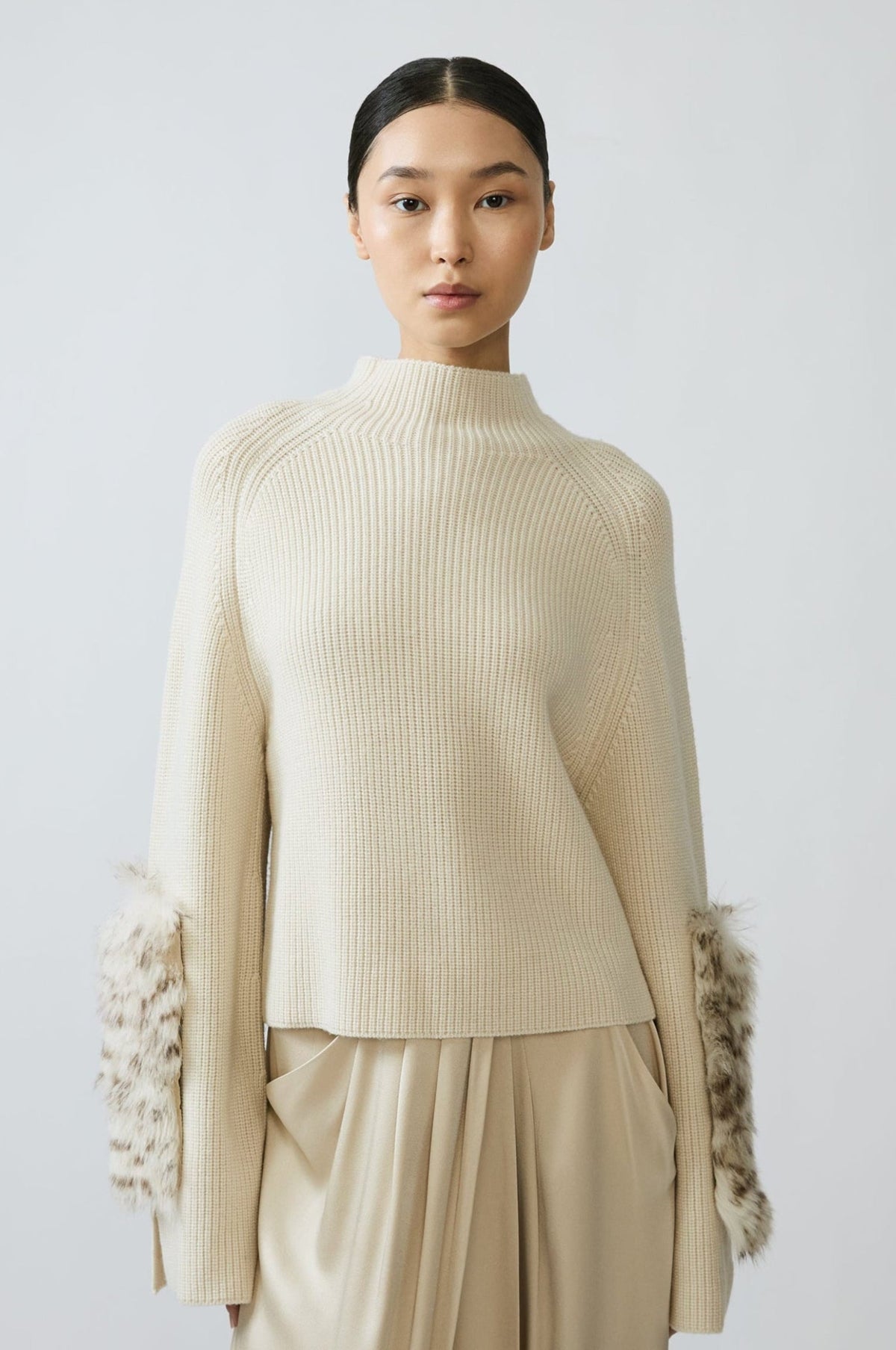 A model wearing a cream ribbed knit mock-neck sweater with textured, fluffy sleeve cuffs and matching cream wide-leg trousers, set against a plain light gray studio background.