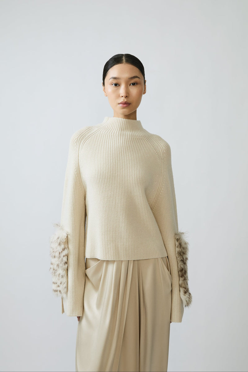 A model wearing a cream ribbed knit mock-neck sweater with textured, fluffy sleeve cuffs and matching cream wide-leg trousers, set against a plain light gray studio background.