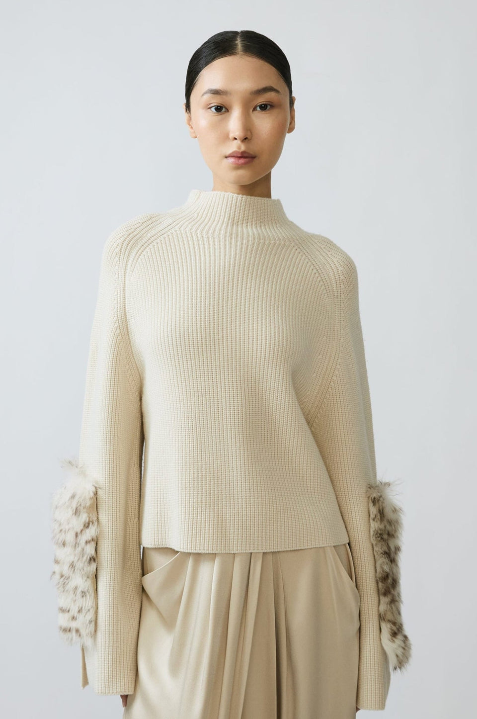 A model wearing a cream ribbed knit mock-neck sweater with textured, fluffy sleeve cuffs and matching cream wide-leg trousers, set against a plain light gray studio background.
