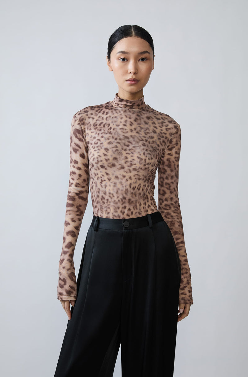 A model wearing a long-sleeve mock neck top with a brown and beige leopard print, paired with black wide-leg trousers, against a light gray studio background.