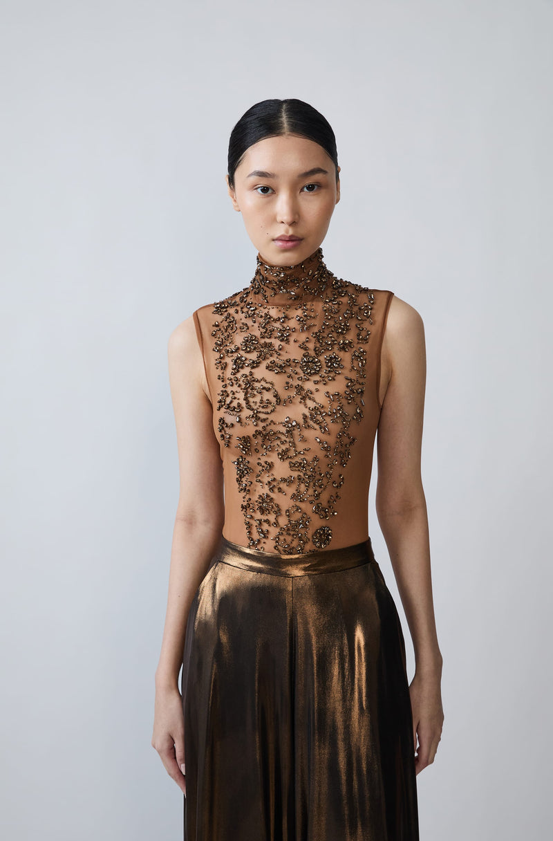 A model wearing a sleeveless high-neck sheer mesh top with intricate black floral embroidery over warm nude skin, paired with a metallic bronze pleated skirt, against a light gray studio background.