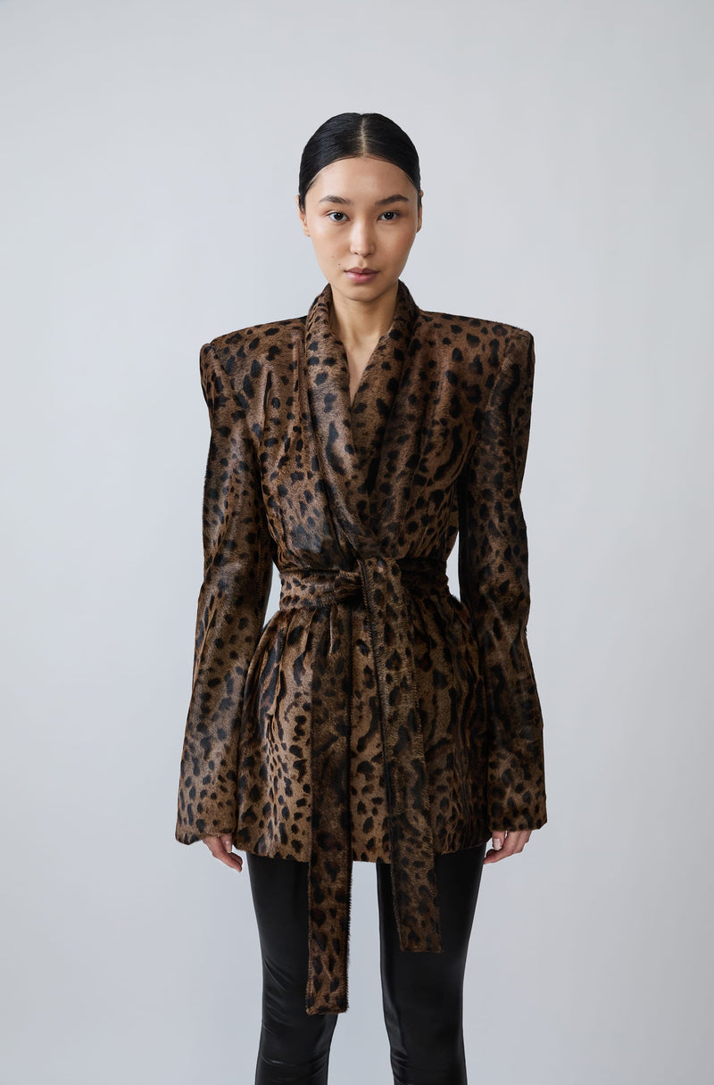 A model wearing a brown and black leopard-print belted wrap jacket with structured shoulders, paired with black pants, against a light gray studio background.